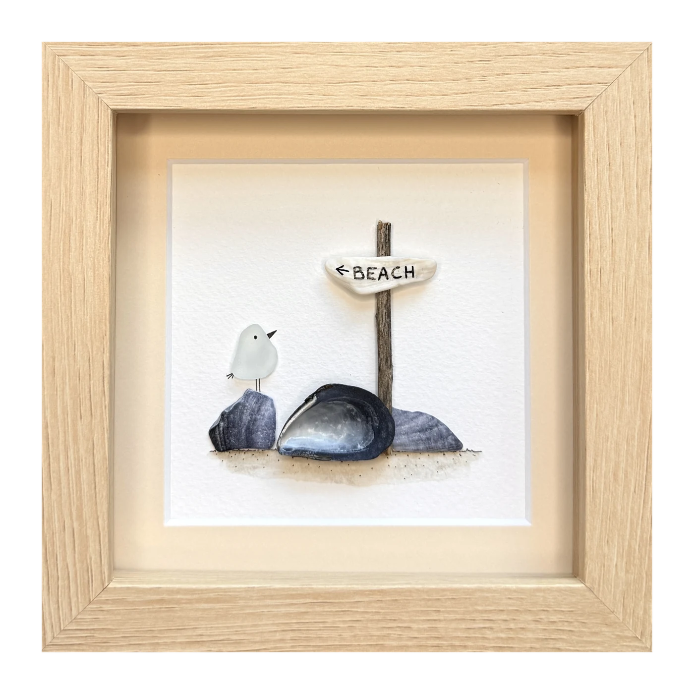 A white sea glass bird on a dark shell beside mussel shells, under a beach sign on a twig
